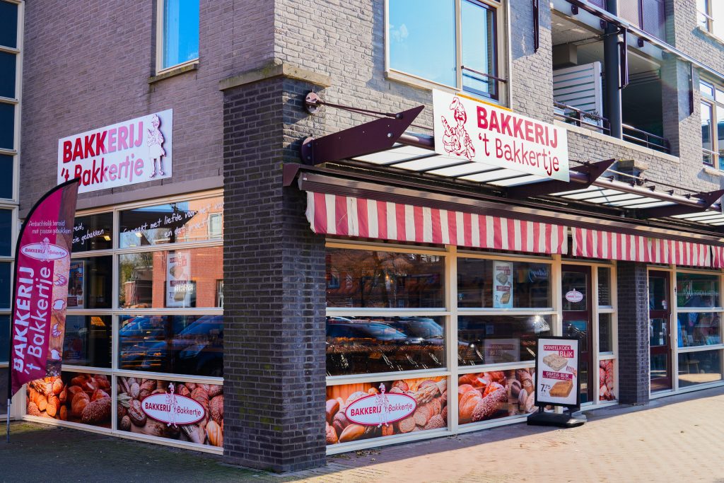 Vooraanzicht van Bakkerij ’t Bakkertje in Mierlo-Hout, Helmond, met een uitnodigende gevel, rood-witte luifel en een sfeervolle etalage vol ambachtelijk brood. Deze centraal gelegen bakkerij aan de Hoofdstraat biedt vers brood, kaneelkoeken en warme worstenbroodjes.